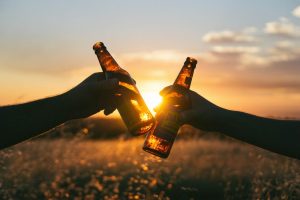 Image of two arms holding beer bottle with sunset and crop in background